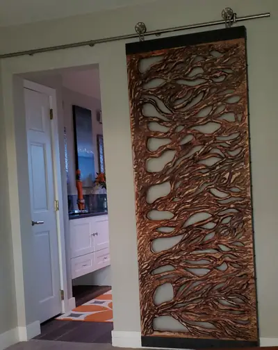A carved wood barn door in a flowing cell pattern, hung on a black rail and backlit by the room behind it.