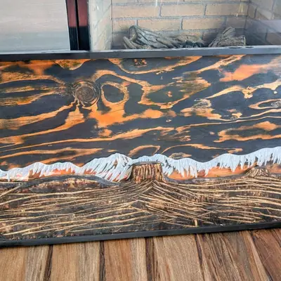 Carved wood wall panel of a snow capped mountain range over layered orange and black scorched wood