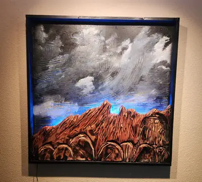 A framed carved wood panel of a mountain range under a heavy storm sky, painted in greys and rust.