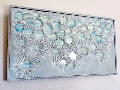 Framed carved wood wall panel covered in overlapping starburst clusters, each centred on a pale blue and green resin disc, hung on a cream wall