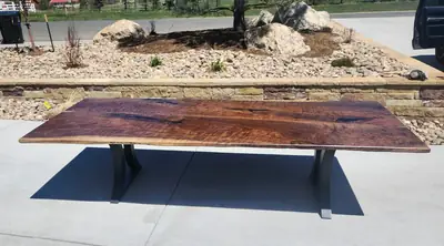 A wide live edge table with a dark poured top on a black steel trestle base, photographed outdoors in daylight.