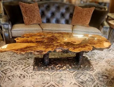 A burl slab coffee table with gold and amber epoxy fill on a sculpted wood base, in front of a tufted leather sofa.