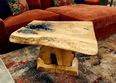 A pale live edge coffee table with a deep blue epoxy pool set into the grain, on a chunky wood pedestal base.