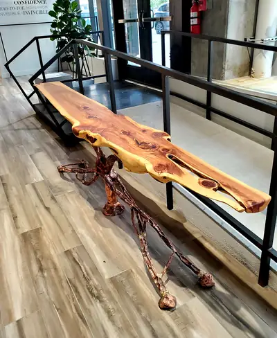 A slim live edge console table resting on a sculpted copper root base, placed against a black railing in an open lobby.