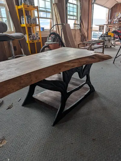 A wide live edge dining table with a soft brown finish, mounted on a sculpted cast iron base in the workshop.