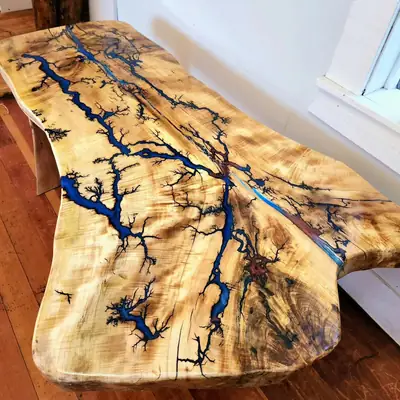 Live edge maple slab table with branching fractal burns filled in deep blue resin running the length of the top, on a wood floor beside a window
