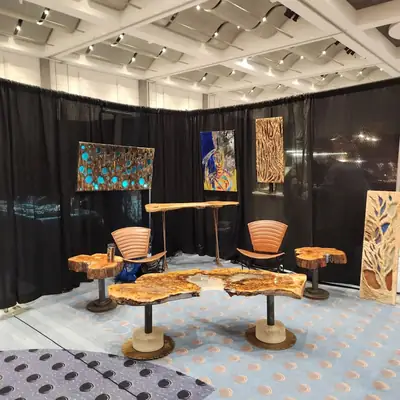 Trade show booth with live edge slab tables on steel pedestals, two leather fan back chairs, and four carved wood wall pieces hung on black drape