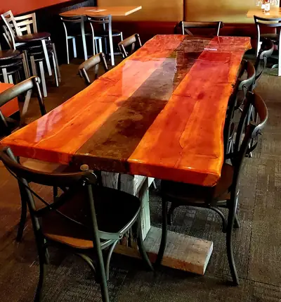 A long live edge dining table in warm red cedar with a mirror gloss finish, set with metal chairs in a bright dining room.