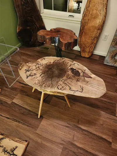 A round live edge table with a dark burned centre and a carved wood base, on a hardwood floor by a window.